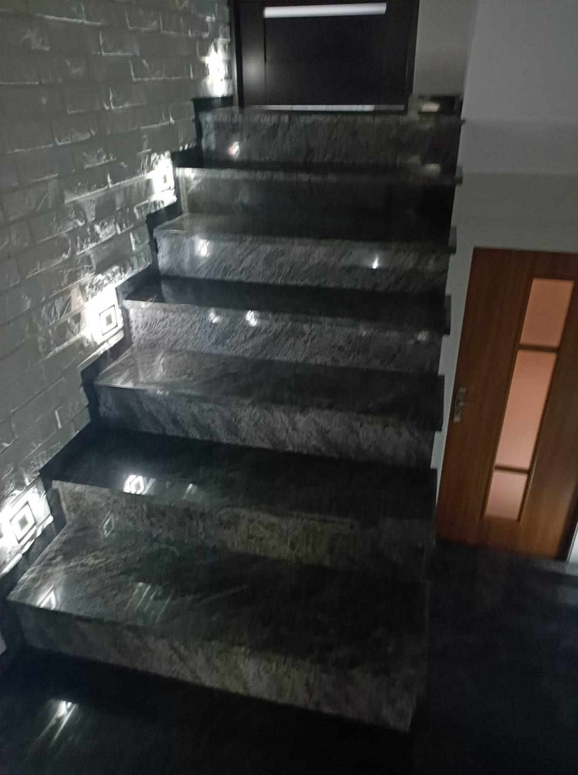 Nowoczesne schody z ciemnego kamienia z subtelnym oświetleniem LED wzdłuż ściany z dekoracyjną fakturą cegły. Widoczne drzwi w tle.