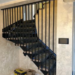 OLstal - Nowoczesne schody z czarną, metalową balustradą w trakcie montażu. Zabezpieczone stopnie, widoczny fragment narzędzi. Ściany w odcieniach beżu i szarości.