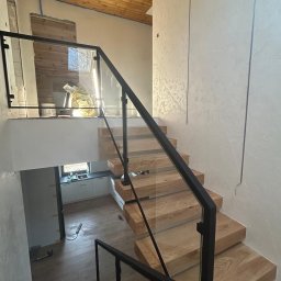 OLstal - Nowoczesne schody drewniane z czarną balustradą szklaną, widok z góry. Minimalistyczny design, jasne drewno, czarne elementy, szklane panele. W tle widoczna kuchnia.