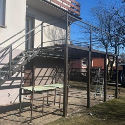 OLstal - Metalowa konstrukcja z schodami na taras w trakcie montażu przy budynku. Widoczne elementy rusztowania i narzędzia. Jasne, słoneczne światło.