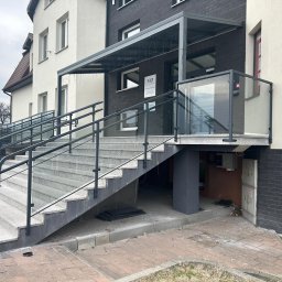 OLstal - Zewnętrzne schody z szarego granitu prowadzące do budynku, zabezpieczone czarnymi poręczami i szklaną balustradą. Nad wejściem zadaszenie z oświetleniem.