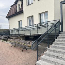 OLstal - Zewnętrzna szklana balustrada z metalowymi elementami, zamontowana przy schodach i podjeździe dla niepełnosprawnych, prowadzących do budynku z jasną elewacją.