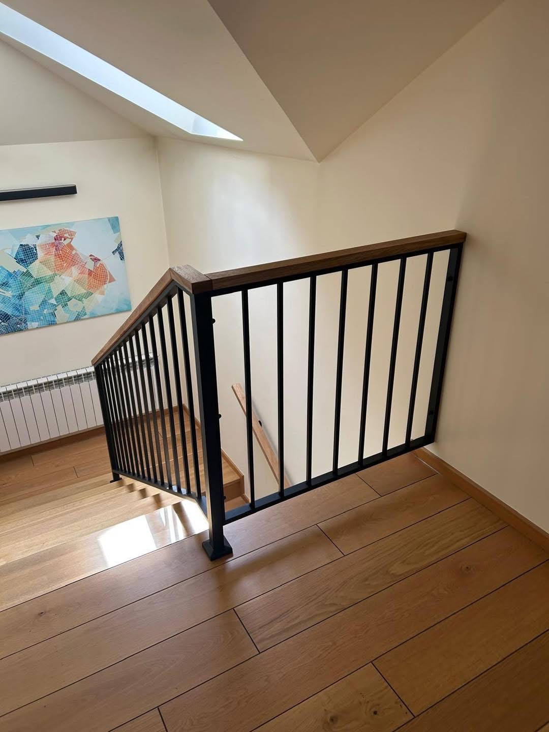 Nowoczesna balustrada schodowa z czarnymi, pionowymi prętami i drewnianym poręczem, oświetlona światłem z okna dachowego. Minimalistyczny design w jasnym wnętrzu.