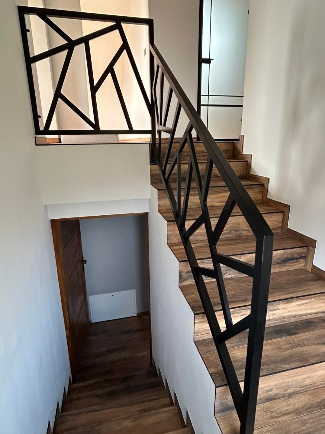 Nowoczesne schody z drewnopodobnymi stopniami i czarną, geometryczną balustradą. Widok z dołu, na białym tle ścian. Otwarta przestrzeń z drzwiami w tle.