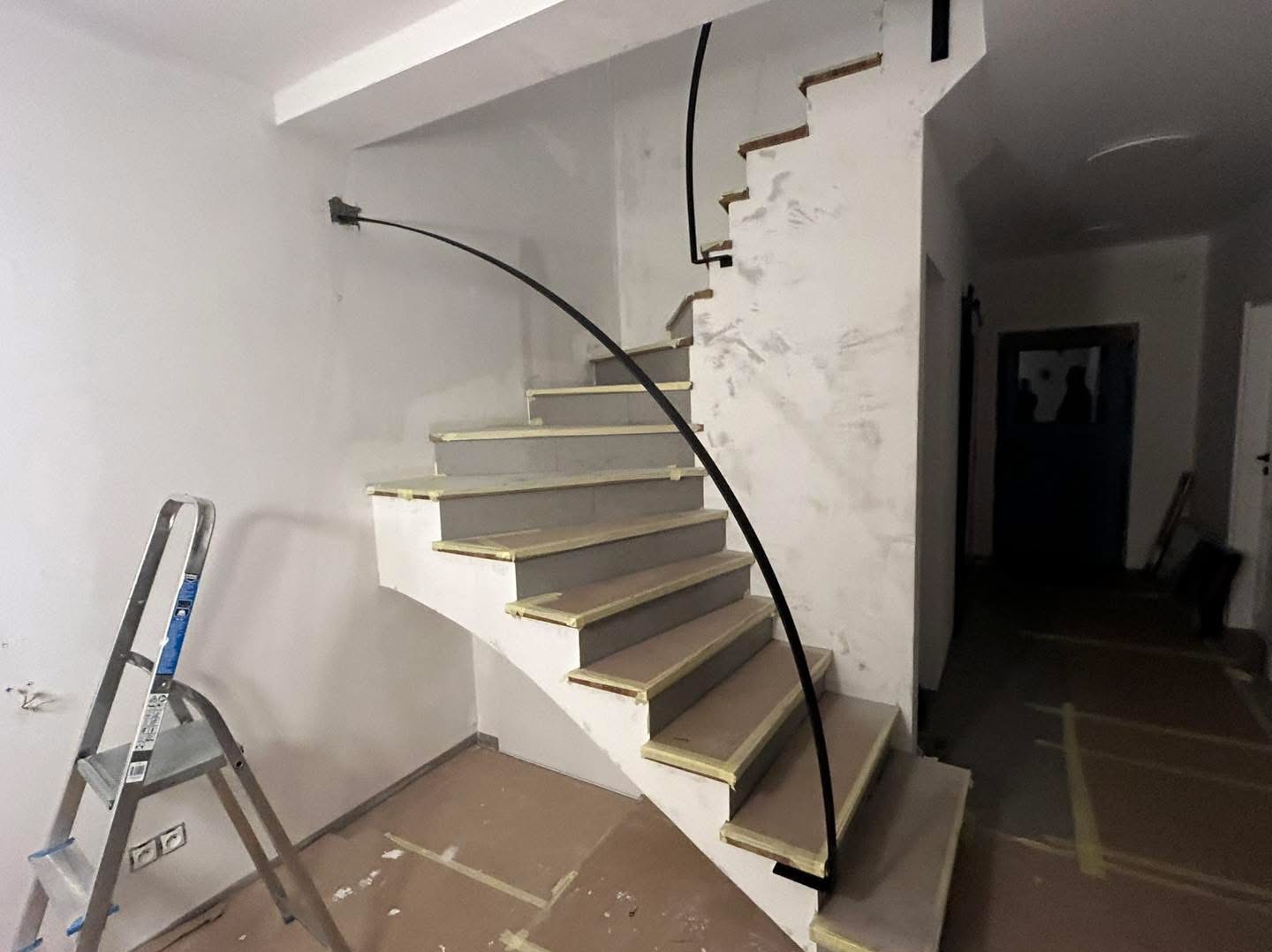 Wnętrze w trakcie wykańczania: spiralne schody z czarną poręczą, zabezpieczone taśmą malarską, drabina oparta o ścianę. Widok na korytarz z drzwiami.