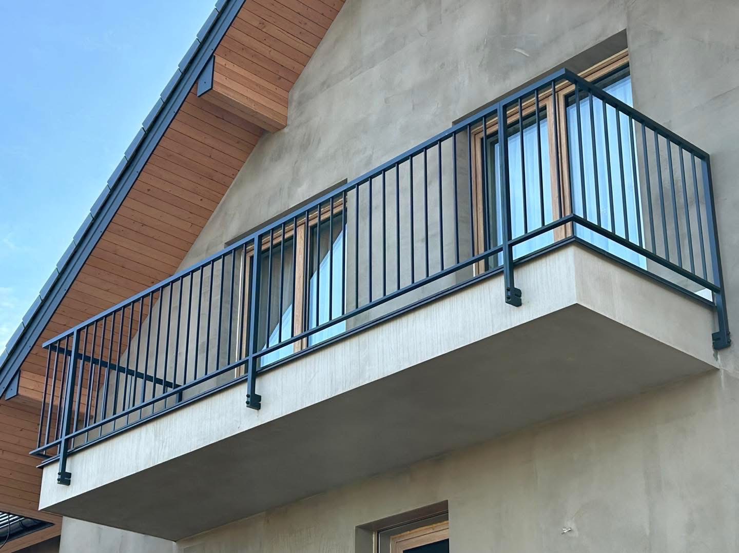 Czarna, metalowa balustrada balkonowa z pionowymi szczeblami, zamontowana na betonowej płycie balkonu w nowoczesnym domu z drewnianym wykończeniem dachu.