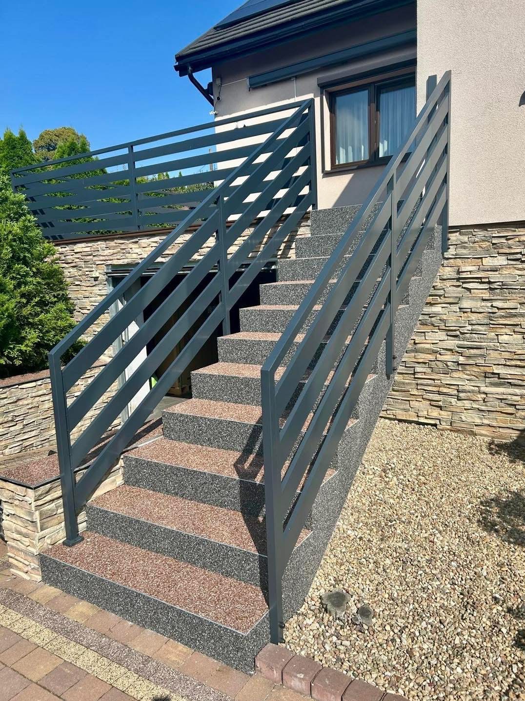 Zewnętrzne schody z szarego metalu z poziomymi balustradami, prowadzące do domu z kamienną elewacją. Widoczne kamienne stopnie i żwirowe podłoże.