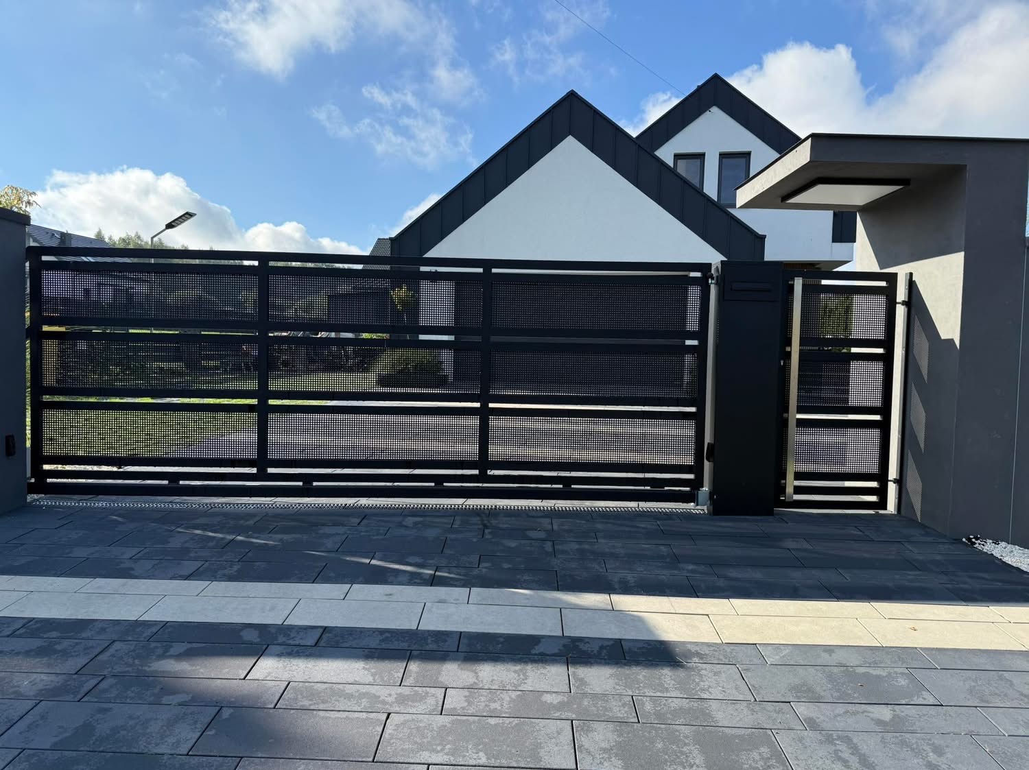 Nowoczesna, czarna brama wjazdowa z perforowanym wypełnieniem na tle domu z dwuspadowym dachem i kostki brukowej w odcieniach szarości. Słoneczny dzień.