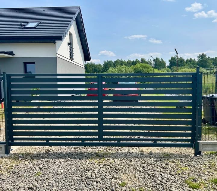 Nowoczesne, poziome ogrodzenie metalowe w kolorze grafitowym, z widocznym fragmentem domu i ogrodu w tle. Minimalistyczny design, równoległe panele, żwirowe podłoże.