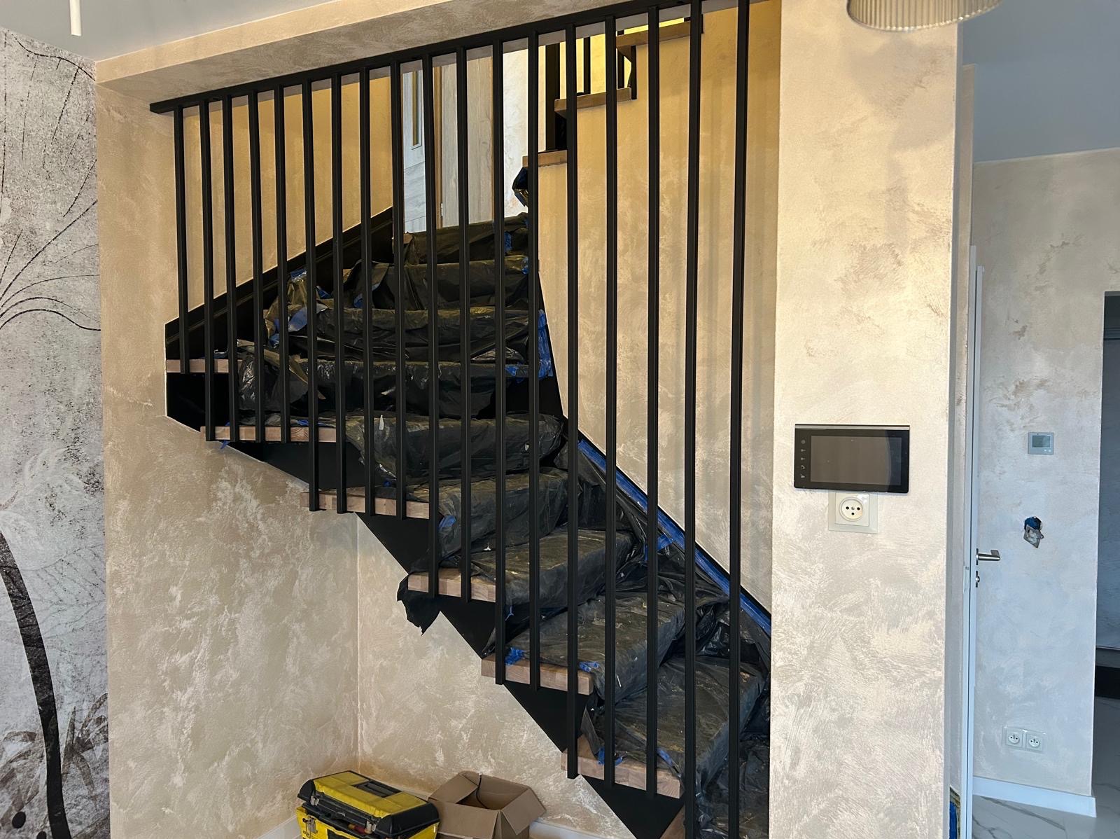 Nowoczesne schody z czarną, metalową balustradą w trakcie montażu. Zabezpieczone stopnie, widoczny fragment narzędzi. Ściany w odcieniach beżu i szarości.