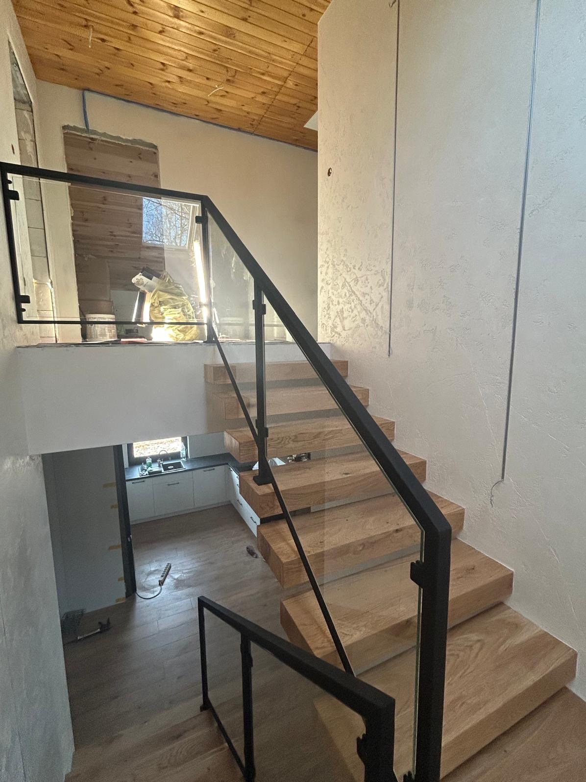 Nowoczesne schody drewniane z czarną balustradą szklaną, widok z góry. Minimalistyczny design, jasne drewno, czarne elementy, szklane panele. W tle widoczna kuchnia.