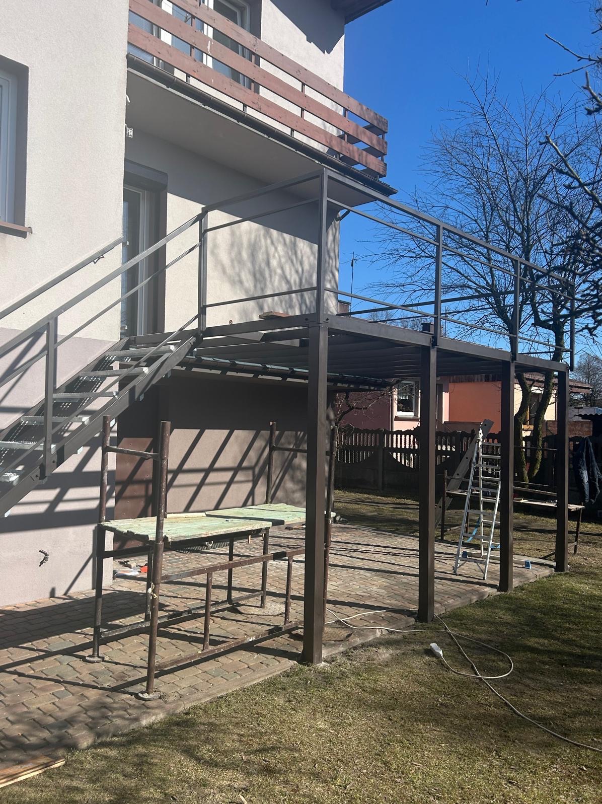 Metalowa konstrukcja z schodami na taras w trakcie montażu przy budynku. Widoczne elementy rusztowania i narzędzia. Jasne, słoneczne światło.