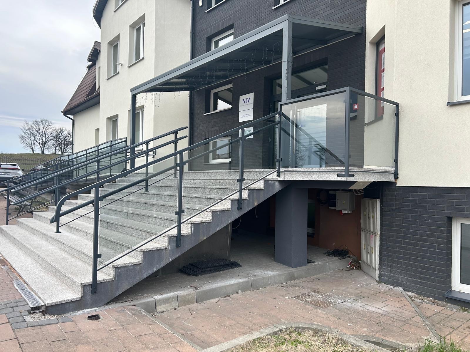 Zewnętrzne schody z szarego granitu prowadzące do budynku, zabezpieczone czarnymi poręczami i szklaną balustradą. Nad wejściem zadaszenie z oświetleniem.