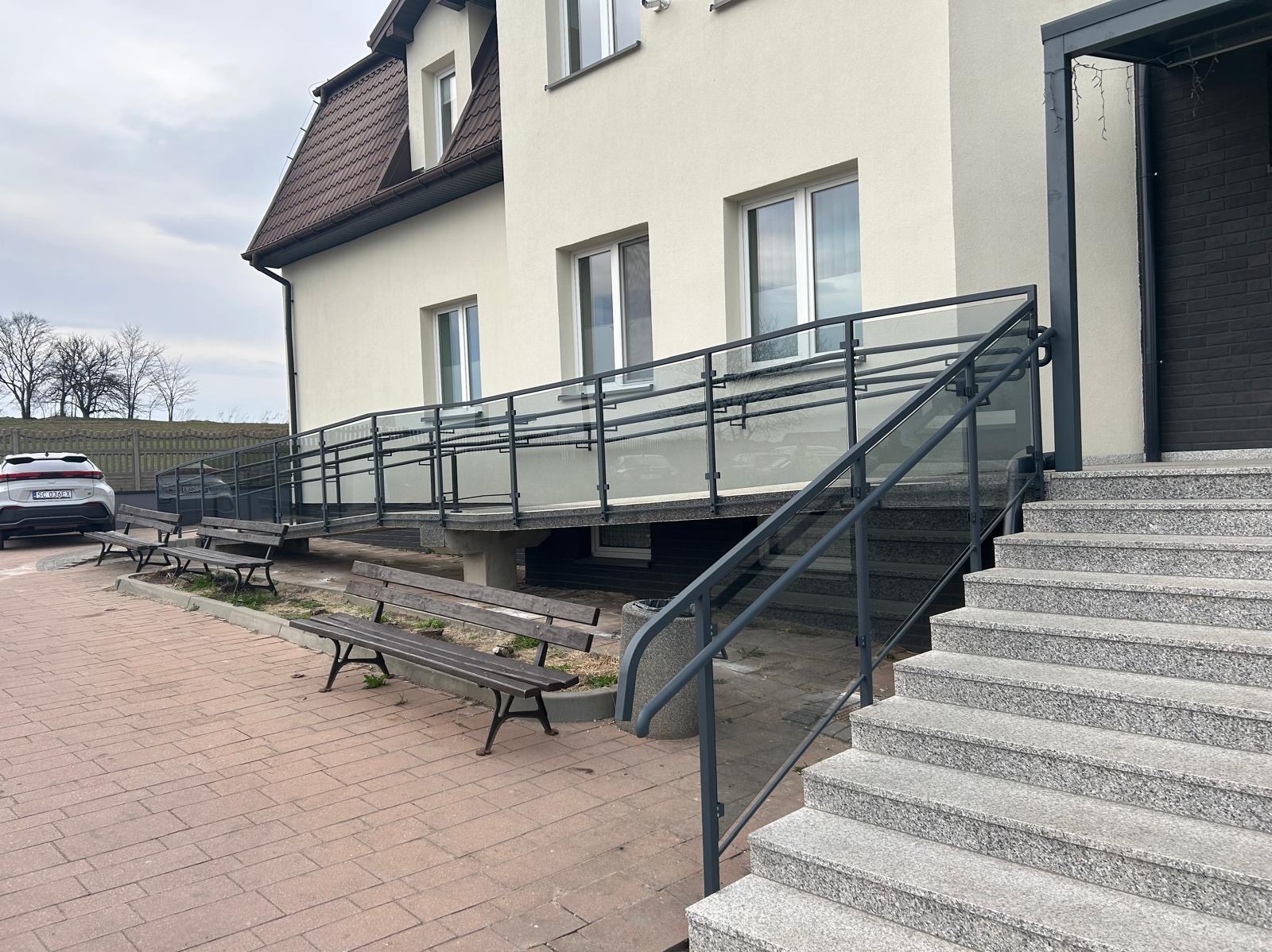 Zewnętrzna szklana balustrada z metalowymi elementami, zamontowana przy schodach i podjeździe dla niepełnosprawnych, prowadzących do budynku z jasną elewacją.