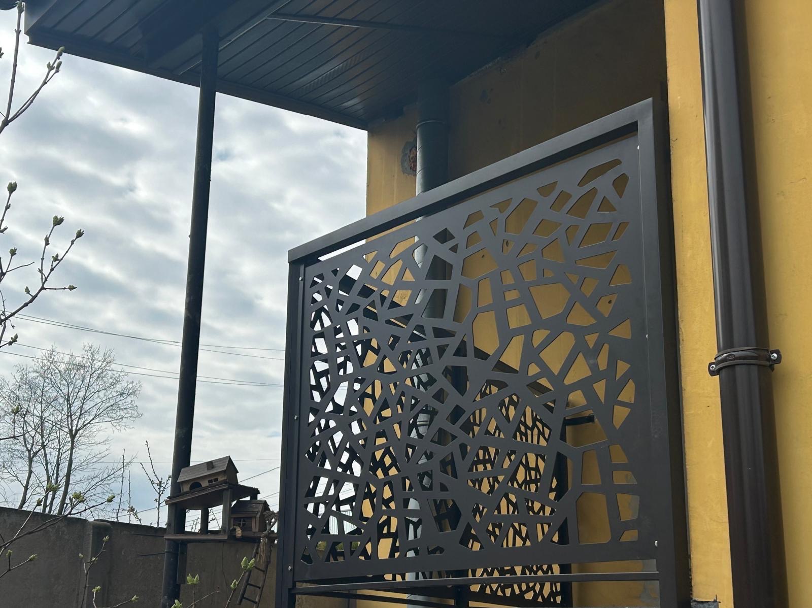 Czarny, metalowy panel ogrodzeniowy z abstrakcyjnym wzorem na tle żółtej ściany budynku i pochmurnego nieba. Widoczny fragment rynny i budka dla ptaków w tle.