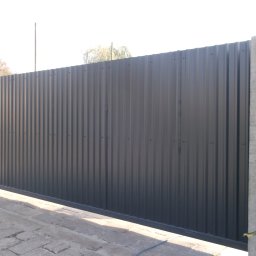 OK Projekt - Nowoczesna, przesuwna brama z szarej blachy trapezowej, zamontowana na betonowych słupkach. Widoczna kostka brukowa i fragment ubrania roboczego na ogrodzeniu w tle.