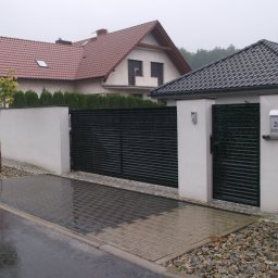 OK Projekt - Nowoczesne, poziome, czarne ogrodzenie panelowe z furtką, biały murek, kostka brukowa na podjeździe, dom z czerwoną dachówką w tle, adres na skrzynce pocztowej.