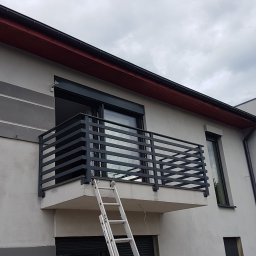 OK Projekt - Nowoczesna, czarna balustrada balkonowa z poziomych profili, zamontowana na jasnym budynku. Aluminiowa drabina oparta o balkon. Pochmurne niebo w tle.