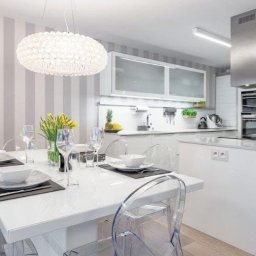 Wiesław Zwoliński - Nowoczesna, jasna kuchnia z białymi meblami i wyspą, akcentowana srebrnym okapem. Stół z przezroczystymi krzesłami i dekoracyjną lampą nad nim. Ściana w szaro-białe pasy.