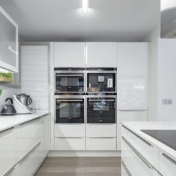 Wiesław Zwoliński - Nowoczesna, biała kuchnia z połyskiem. Zabudowa kuchenna z wbudowanymi piekarnikami, minimalistyczny okap, akcesoria kuchenne i rośliny na blacie. Stonowana kolorystyka.