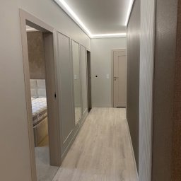 Wiesław Zwoliński - Korytarz w neutralnych barwach z oświetleniem LED podsufitowym, panelami ściennymi z lustrami i widokiem na sypialnię. Jasna podłoga i drzwi w odcieniach beżu.