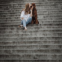 Fotografia Filomeny Aleksandra Dąbrowska - Kobieta w białej koszuli i jeansach siedzi na schodach, całując rudego psa rasy seter irlandzki. Stonowana kolorystyka, naturalne światło.