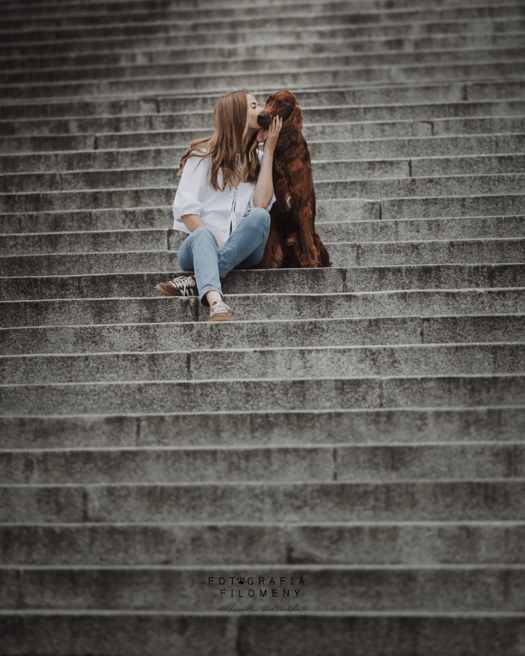 Kobieta w białej koszuli i jeansach siedzi na schodach, całując rudego psa rasy seter irlandzki. Stonowana kolorystyka, naturalne światło.