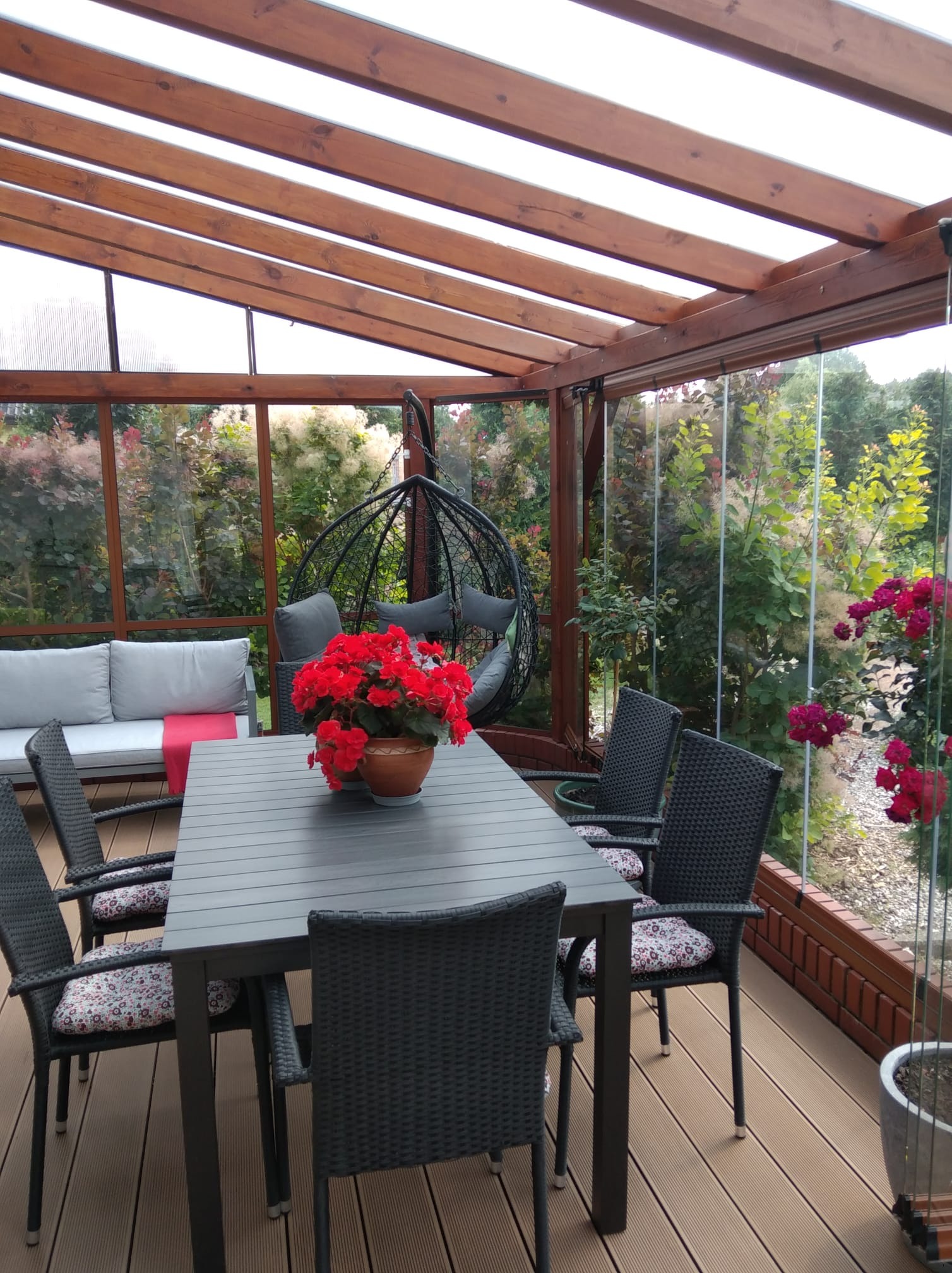 Zadaszony taras z drewnianą konstrukcją i szklanymi ścianami. Wnętrze wyposażone w stół z krzesłami, sofę i wiszący fotel. Na stole stoi doniczka z czerwonymi kwiatami.