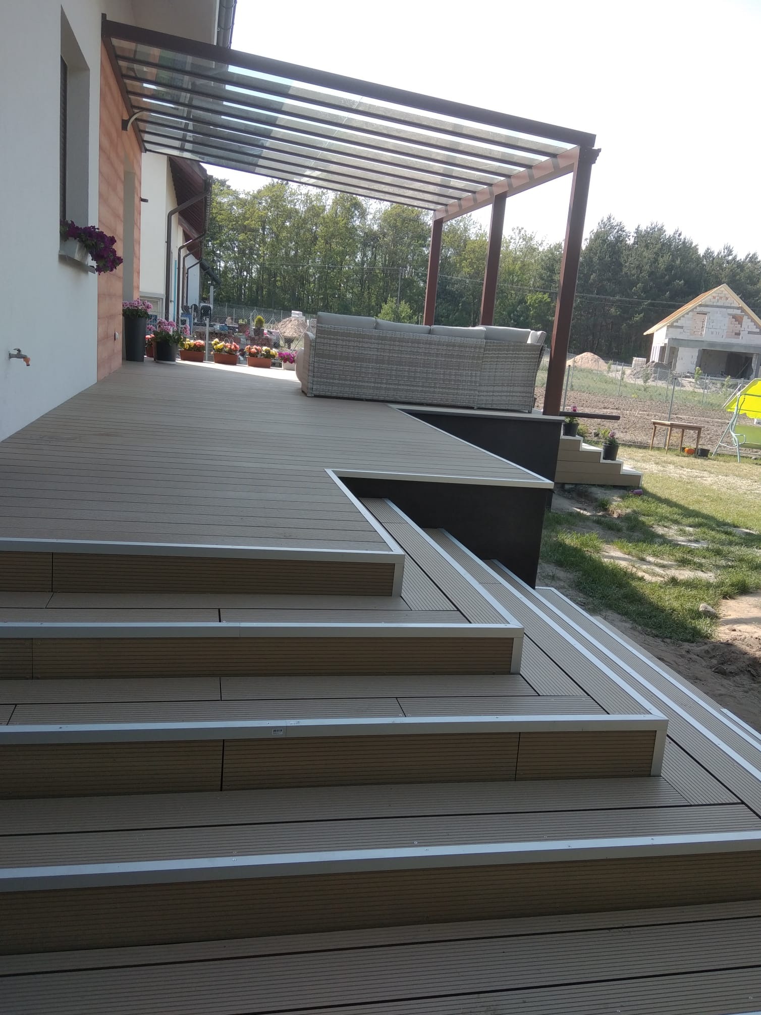 Nowoczesny taras z kompozytu w odcieniach brązu, zadaszony pergolą. Widoczne schody z srebrnymi listwami, sofa ogrodowa i kwiaty w donicach. W tle dom w budowie.