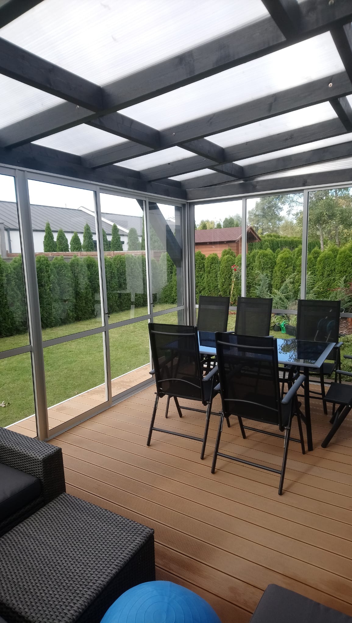 Zadaszony taras z meblami ogrodowymi i widokiem na zadbany ogród. Konstrukcja z szarych belek i przezroczystego dachu. Podłoga z desek w ciepłym odcieniu brązu.