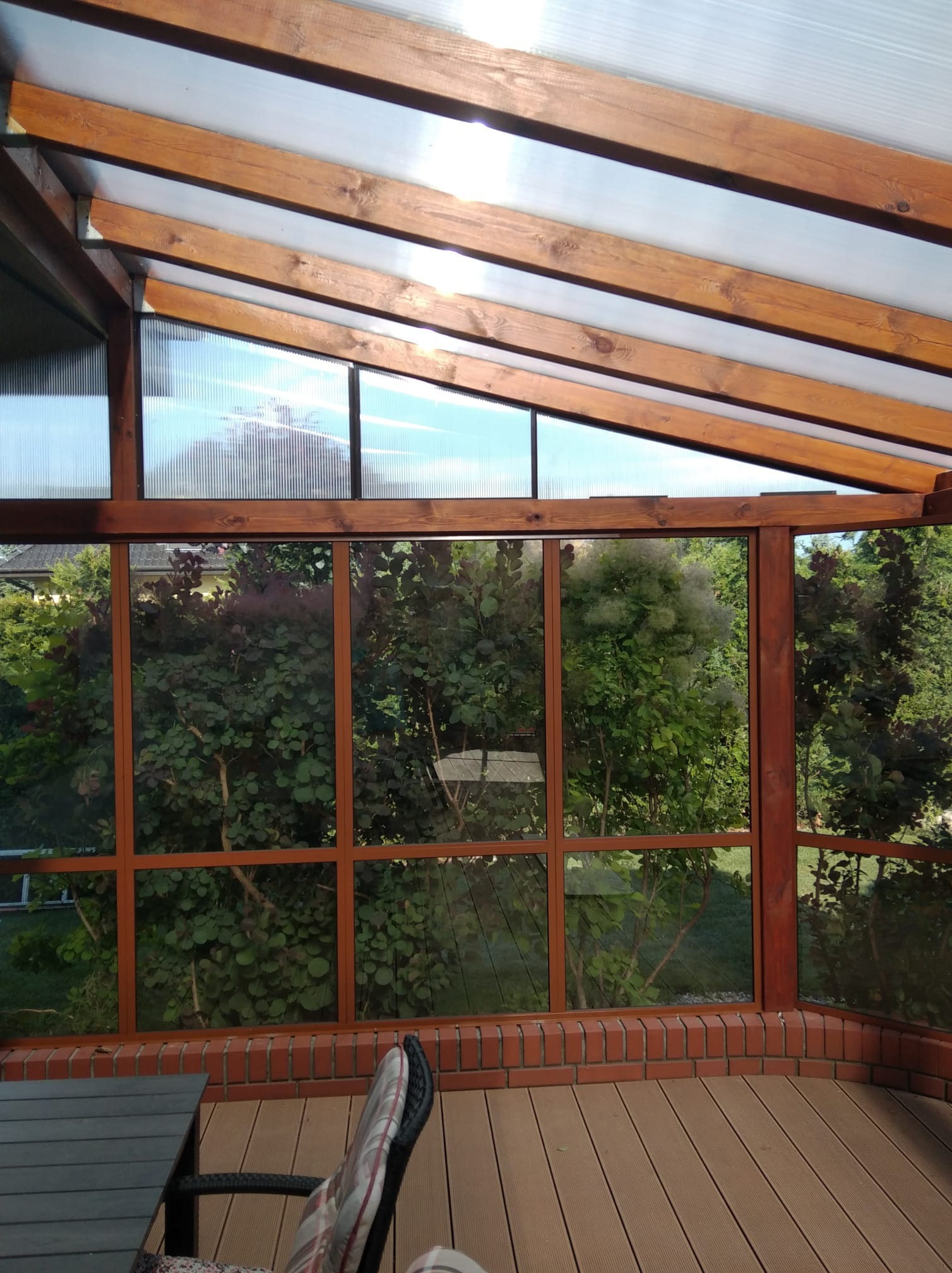 Zadaszony balkon z drewnianą konstrukcją i widokiem na zieleń. Na podłodze deski, częściowo widoczne krzesło i stół. Przeszklenia z widokiem na ogród.