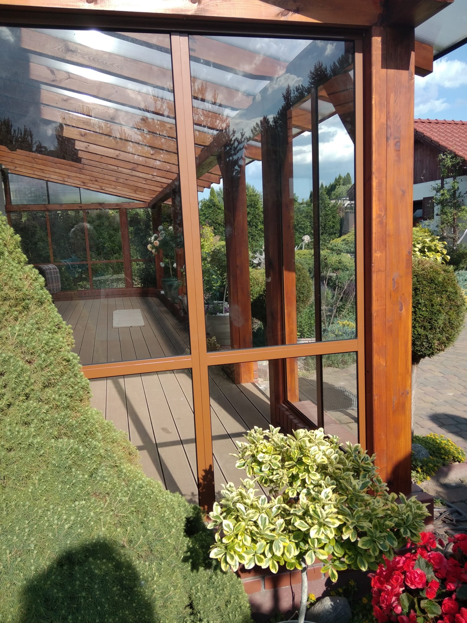 Zadaszona weranda z drewnianą konstrukcją i przeszklonymi ścianami, widok na ogród. Podłoga z desek kompozytowych, rośliny ozdobne na pierwszym planie.