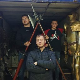 Szymon Piałucha - Transport scenografii: Trzej mężczyźni pozują wewnątrz niebieskiego busa z dwoma zbrojami rycerskimi (czarną i złotą) oraz halabardą. W tle widoczny budynek i zaparkowany samochód.