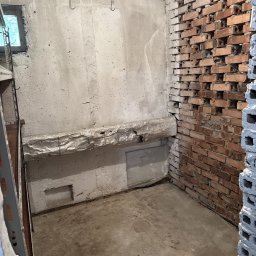 Pustostan - Pusty, surowy piwniczny pokój z odsłoniętą cegłą, betonowymi ścianami i podłogą. Widoczne haki na ścianie, rura owinięta folią i fragment regału po lewej.