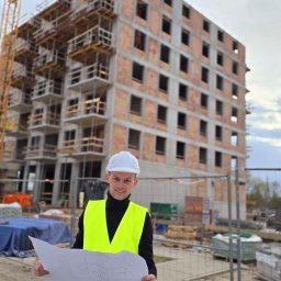 InżynierControl Michał Chomyszyn - Inżynier w kasku i kamizelce odblaskowej trzyma plan na tle budynku w trakcie budowy z widocznym żurawiem. Uśmiechnięty, patrzy w obiektyw.