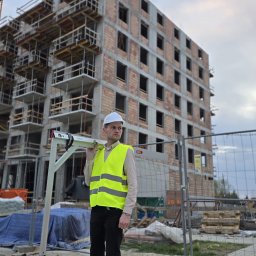 InżynierControl Michał Chomyszyn - Inżynier w kasku i kamizelce odblaskowej z poziomicą na tle budynku w trakcie budowy. Widoczne rusztowania i elementy konstrukcyjne.