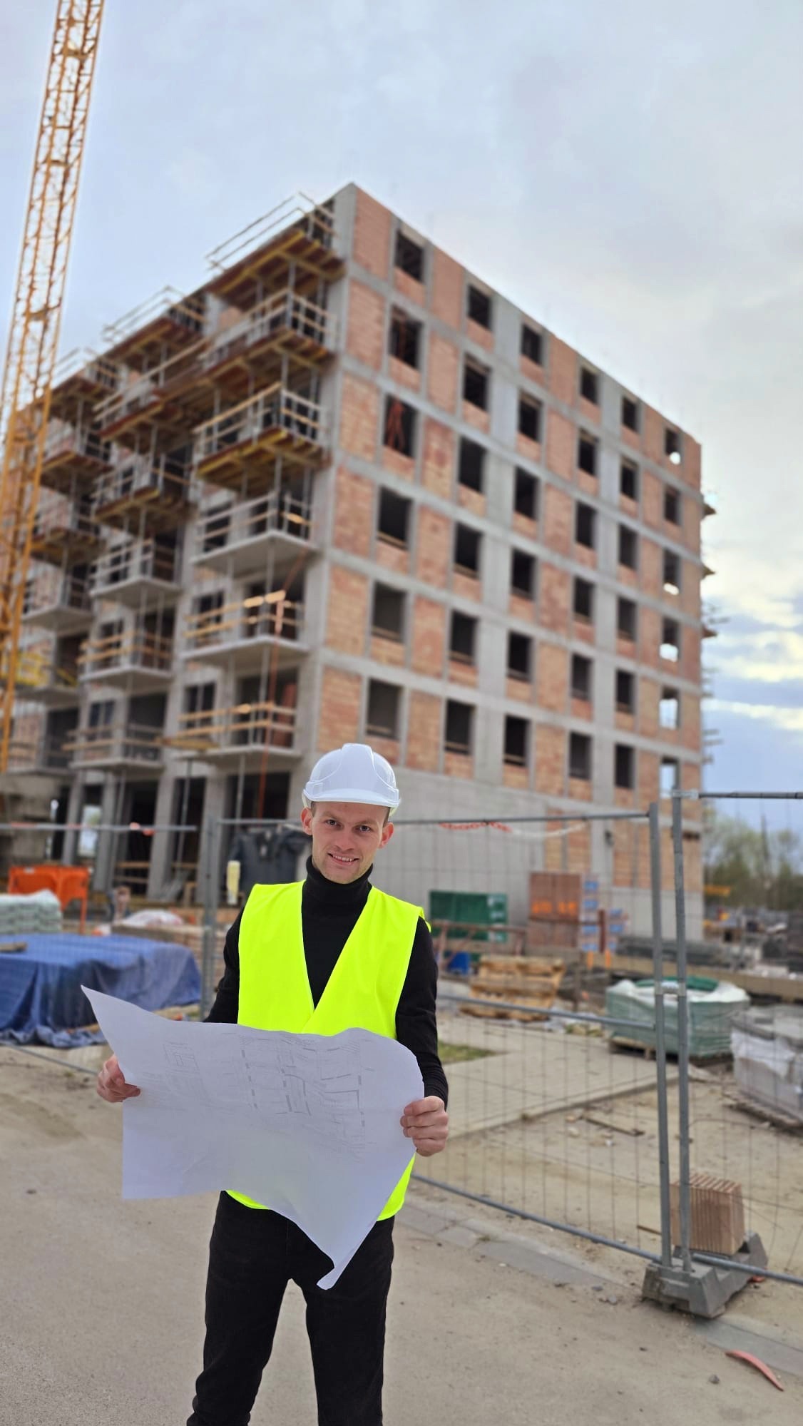 Inżynier w kasku i kamizelce odblaskowej trzyma plan na tle budynku w trakcie budowy z widocznym żurawiem. Uśmiechnięty, patrzy w obiektyw.