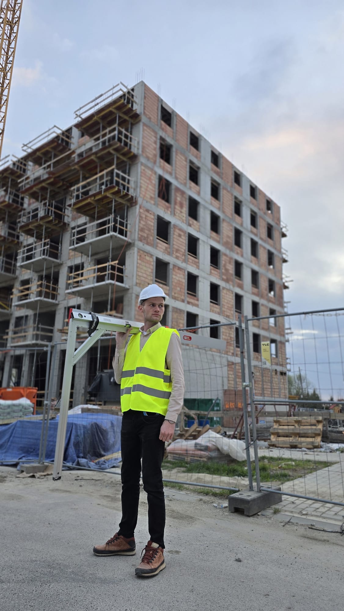 Inżynier w kasku i kamizelce odblaskowej z poziomicą na tle budynku w trakcie budowy. Widoczne rusztowania i elementy konstrukcyjne.