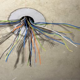 Krzysztof Sobina - Wiązka kolorowych kabli elektrycznych wychodzących z okrągłego otworu w szarej ścianie, z widocznymi odręcznymi notatkami na ścianie.