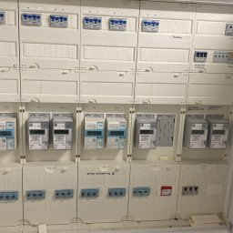 Krzysztof Sobina - Otwarta, biała szafa rozdzielcza z licznikami i zabezpieczeniami, widoczne opisy w języku niemieckim. Instalacja elektryczna wewnątrz budynku.
