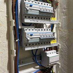 Krzysztof Sobina - Rozdzielnica elektryczna Hager z zainstalowanymi bezpiecznikami i okablowaniem w kolorach: niebieskim, brązowym i żółto-zielonym. Widoczne elementy montażowe i oznaczenia.