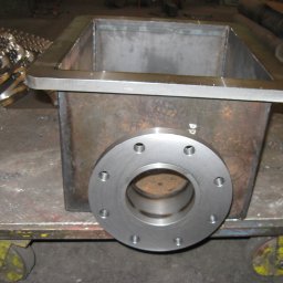 PPUH ZMETIS-BIS MAREK SOSNOWSKI - Spawana konstrukcja ze stali nierdzewnej z kołnierzem, umieszczona na wózku warsztatowym. Widoczne ślady obróbki i spawania. W tle elementy metalowe.