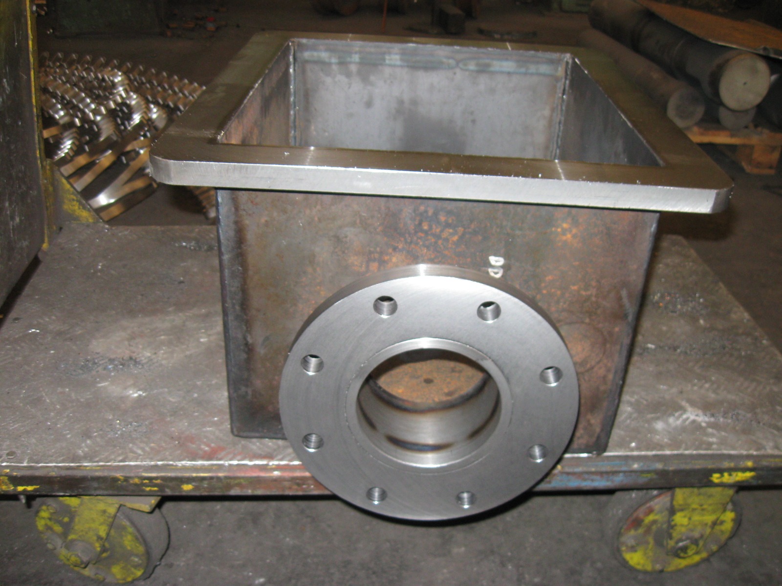 Spawana konstrukcja ze stali nierdzewnej z kołnierzem, umieszczona na wózku warsztatowym. Widoczne ślady obróbki i spawania. W tle elementy metalowe.