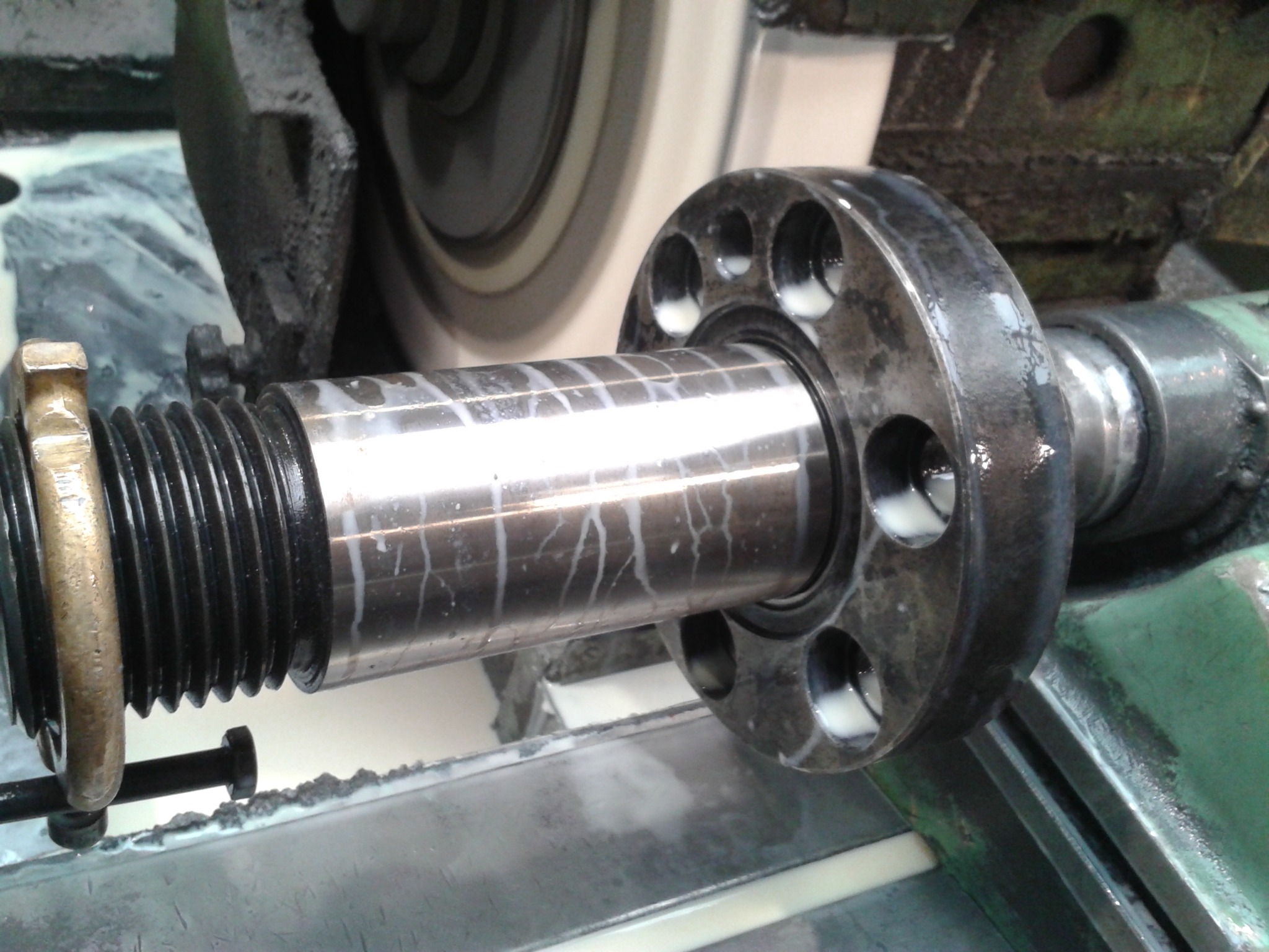 Obróbka tokarska metalowego wału z kołnierzem na tokarce. Widoczne ślady chłodziwa i gwintowany koniec wału.