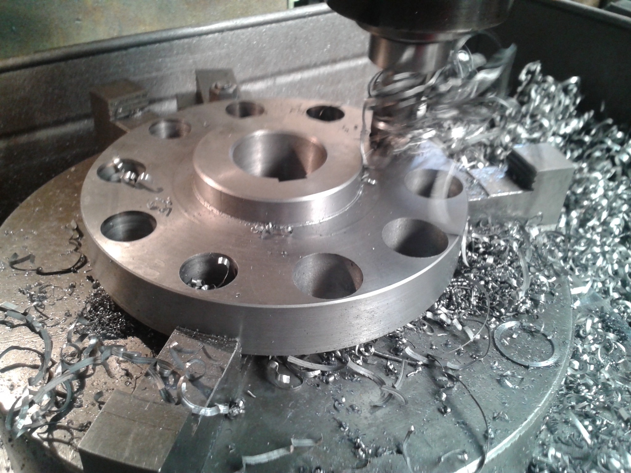 Obróbka metalu: wiertło frezuje okrągły element z otworami, wióry metalowe wokół. Widoczne uchwyty mocujące. Szary odcień metalu dominuje w kadrze.