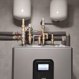 OZESYSTEM - Szara pompa ciepła Thermia z białymi zbiornikami wyrównawczymi i izolowanymi rurami w pomieszczeniu o szarych ścianach. Widoczny ekran z parametrami pracy.