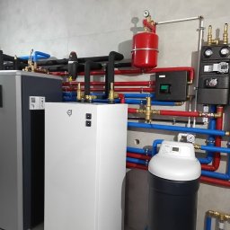 OZESYSTEM - Kompletna instalacja grzewcza z pompą ciepła, zbiornikiem i rozdzielaczami, rury w izolacji czerwonej i niebieskiej, manometry, nowoczesny design, szare płytki na ścianie.