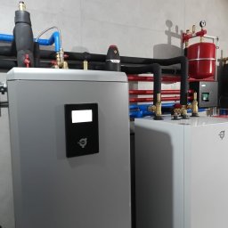OZESYSTEM - Złożona instalacja z pompą ciepła Thermia w Płońsku. Widoczne rury w izolacji, zbiornik wyrównawczy i sterownik. Szara ściana w tle.
