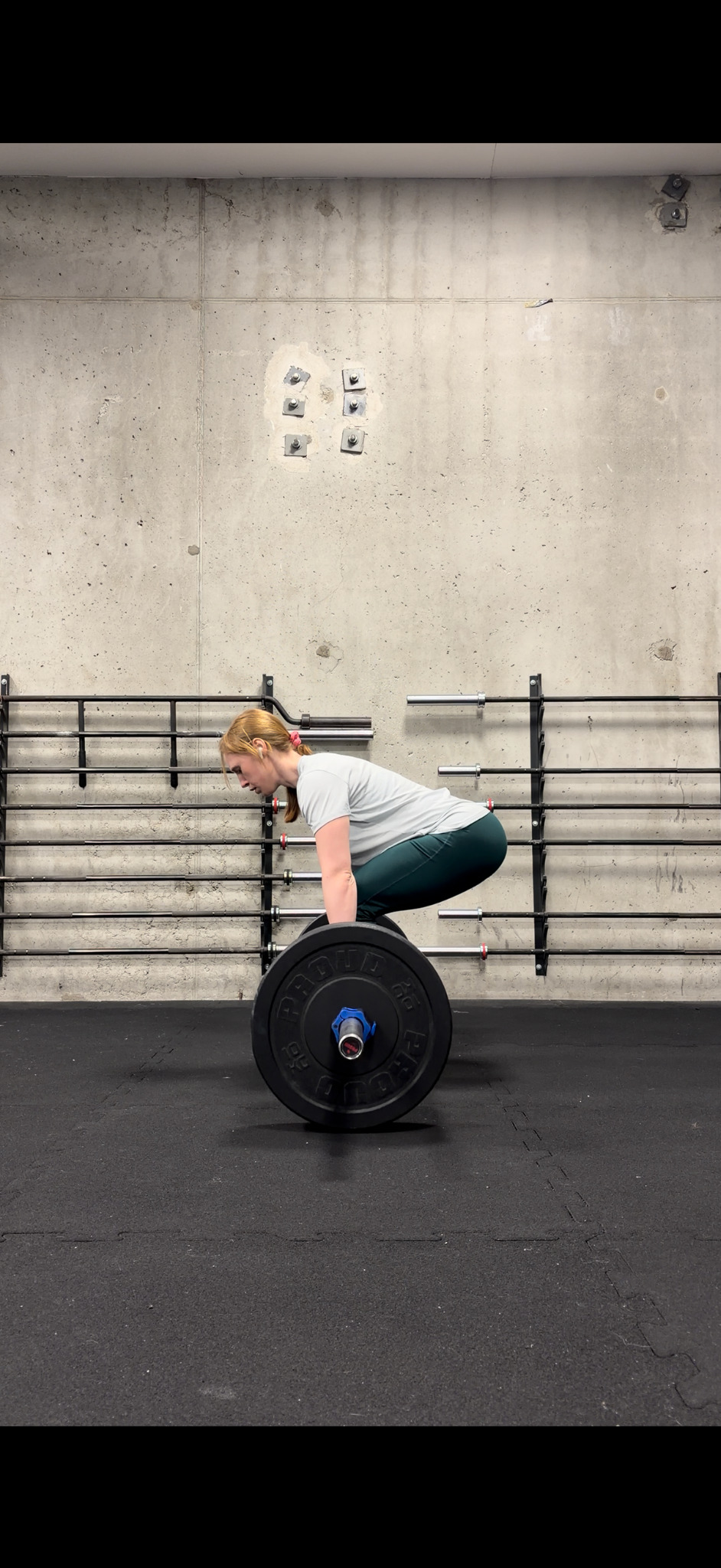 Kobieta w szarym t-shircie wykonuje martwy ciąg ze sztangą w siłowni. Widok z przodu, betonowa ściana w tle, czarna podłoga z gumy.