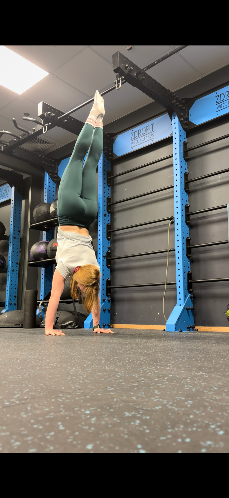 Kobieta w zielonych legginsach wykonuje stójkę przy drabinkach w siłowni Zdrofit. Widoczne piłki lekarskie i elementy wyposażenia siłowni.